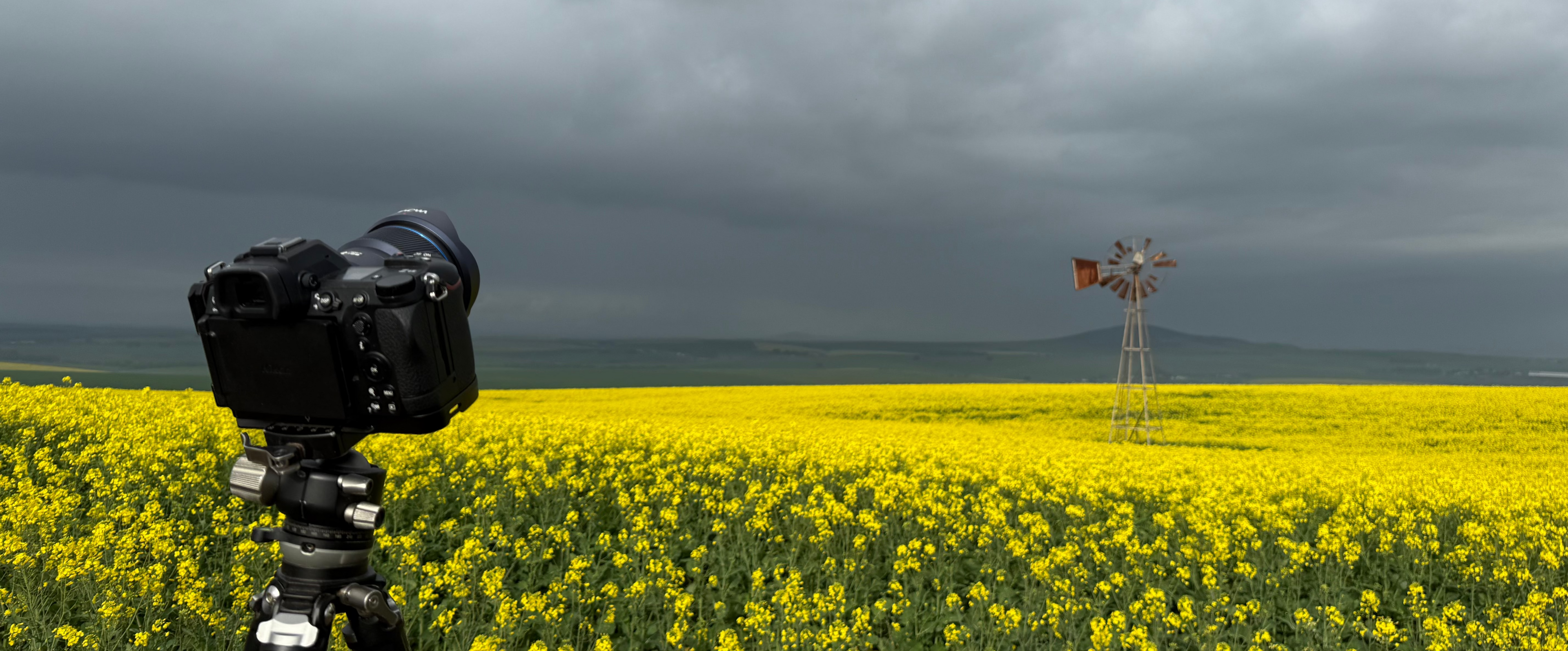 Camera on a tripod pointing towards a yellow field with a windmill under a cloudy sky.