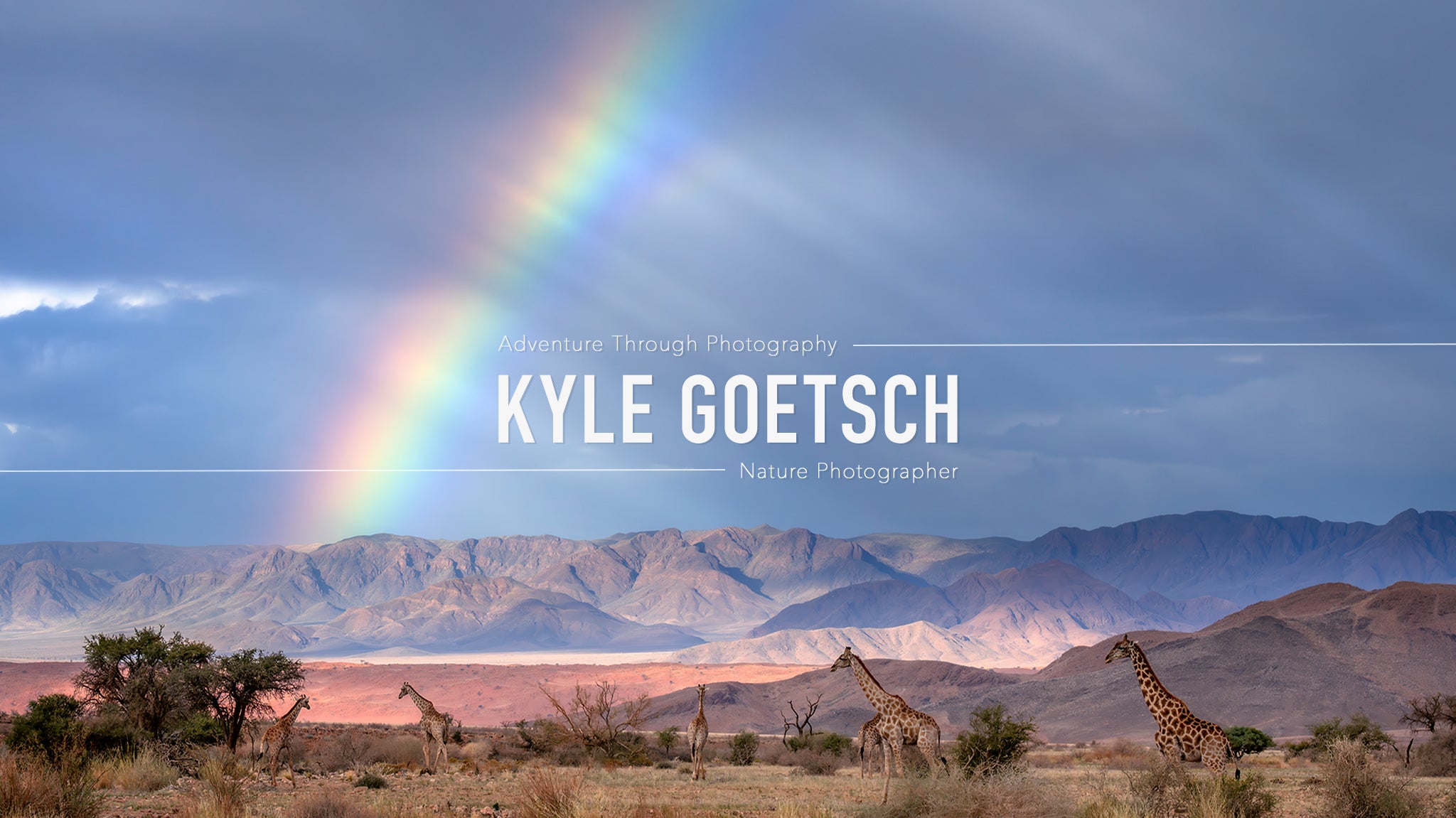 Giraffes in a desert landscape with a rainbow and mountains, credited to Kyle Goetsch.