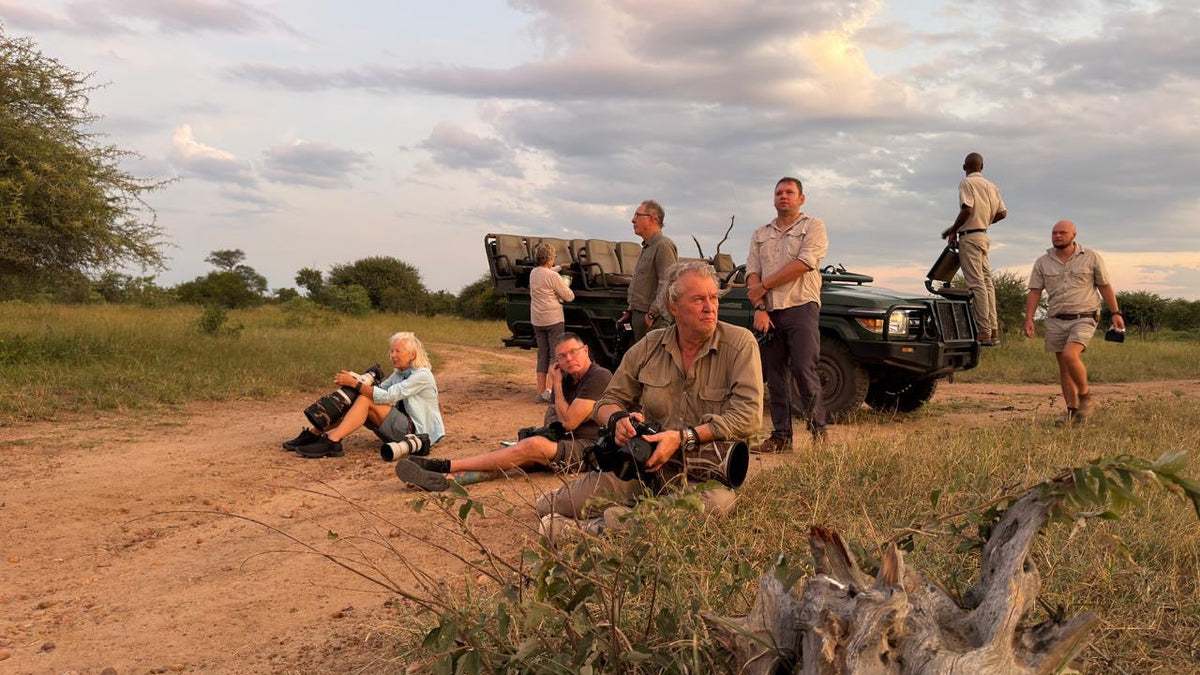 A safari group of people relaxes and chats on a dirt road next to a green safari vehicle in a grassy savanna landscape under a cloudy sky.