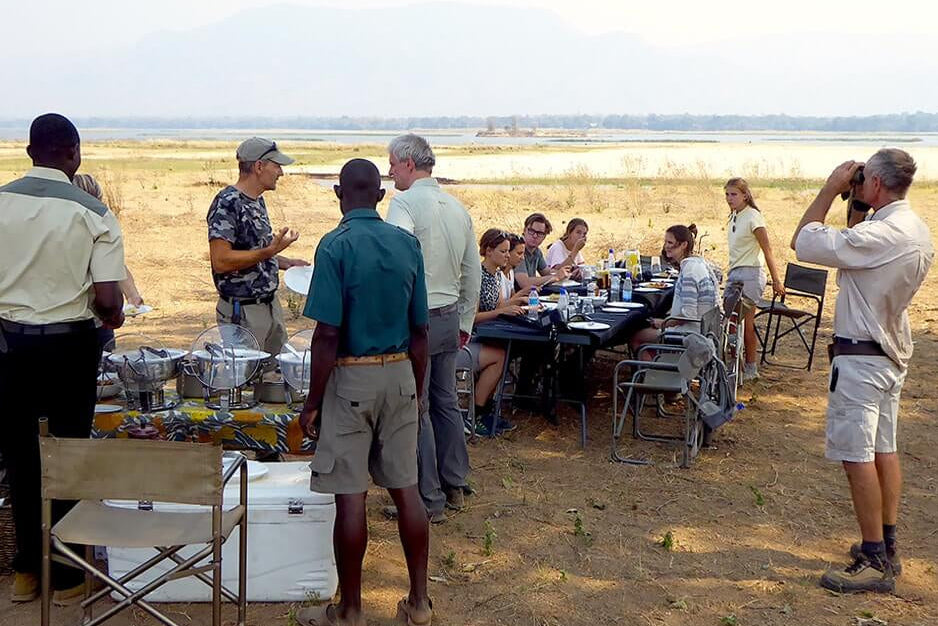 Group of people gathered around a table in an open field with chairs and a cooler.